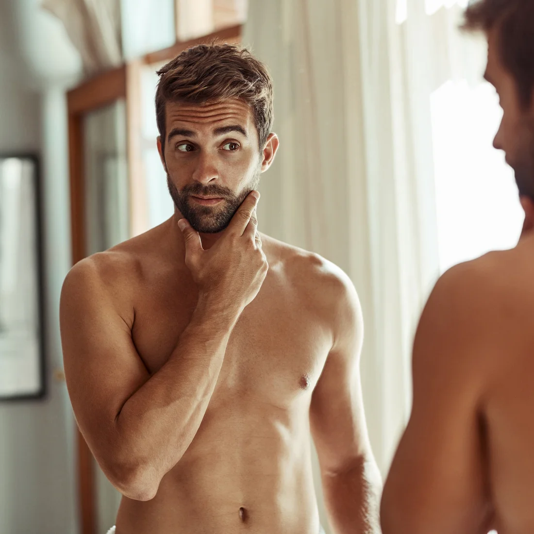 Bare-chested man touching jaw and looking in the mirror